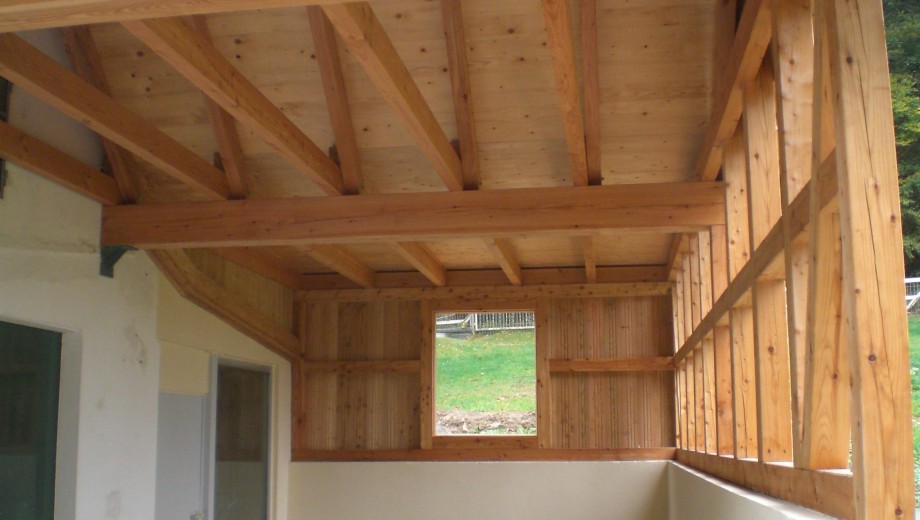 Teilweise überdachte Holzkonstruktion mit Fenster, umgeben von hellen Wänden und natürlicher Umgebung.