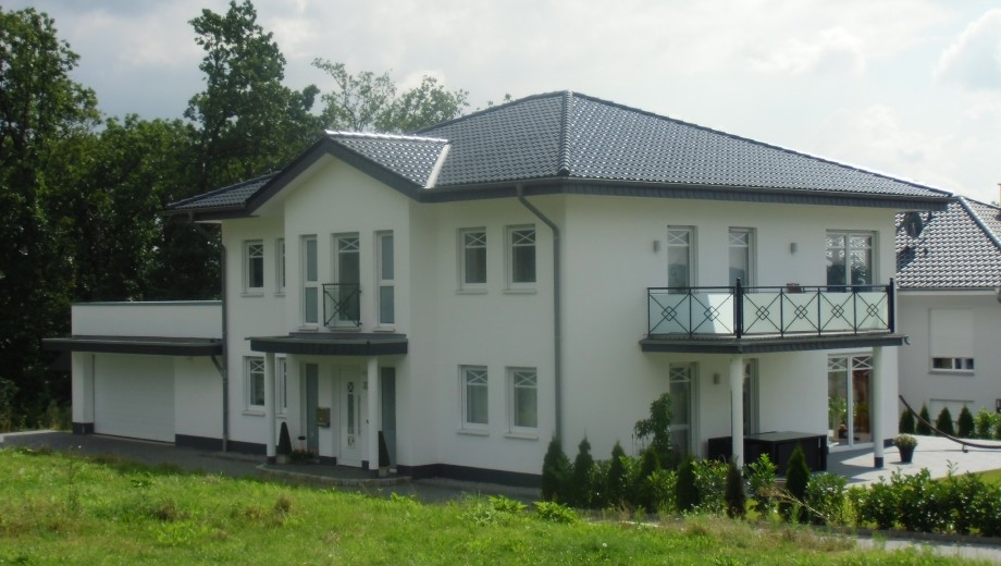 Modernes zweistöckiges Einfamilienhaus mit weißer Fassade, dunklem Dach und grünem Rasen im Vordergrund.