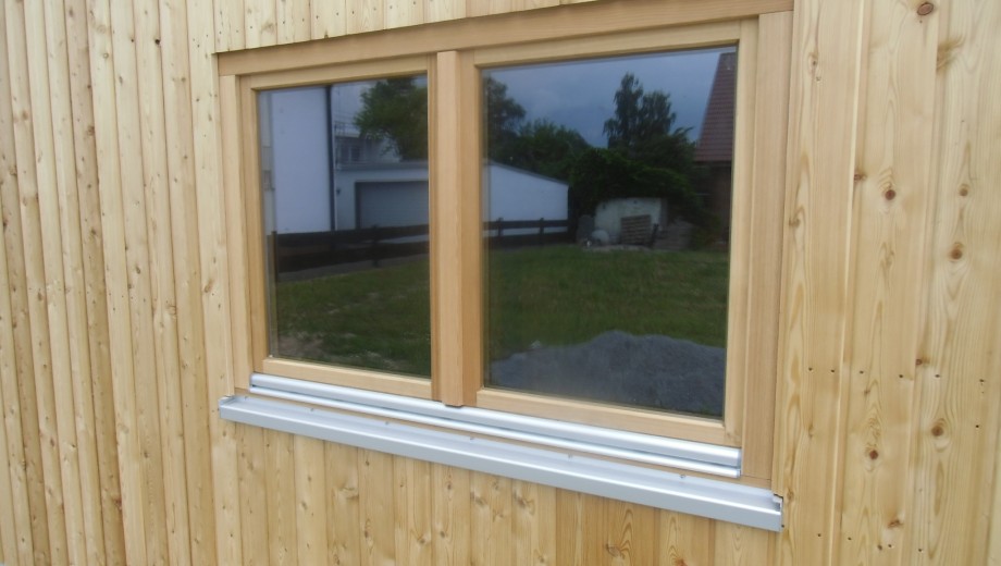 Holzfassade mit einem doppelflügeligen Fenster, das Bäume und ein Gebäude reflektiert.