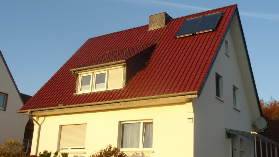 Einfamilienhaus mit rotem Ziegeldach und Solarpanel, cremeweiße Fassade, ruhige Wohngegend.
