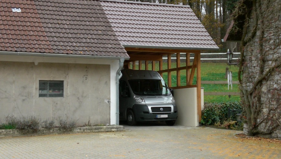 Ein grauer Lieferwagen steht in einem Carport neben einem Gebäude mit Ziegeldach in ländlicher Umgebung.
