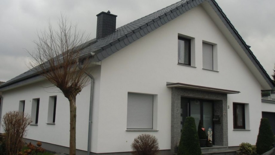 Modernes Einfamilienhaus mit weißer Fassade, dunklem Dach und gepflegtem Garten unter grauem Himmel.