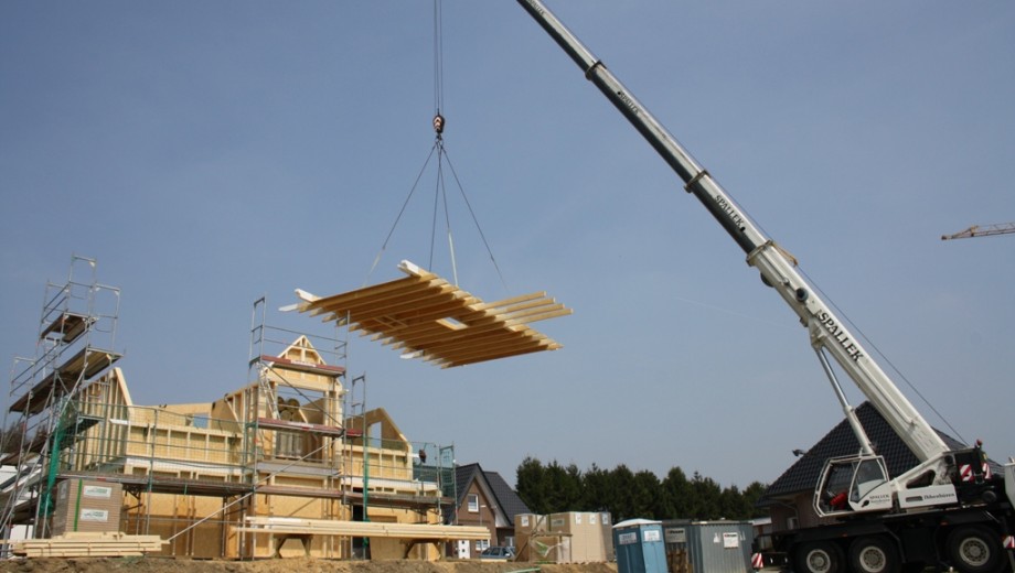 Ein Kran hebt Holzelemente zu einem im Bau befindlichen Gebäude mit Gerüst unter klarem Himmel.