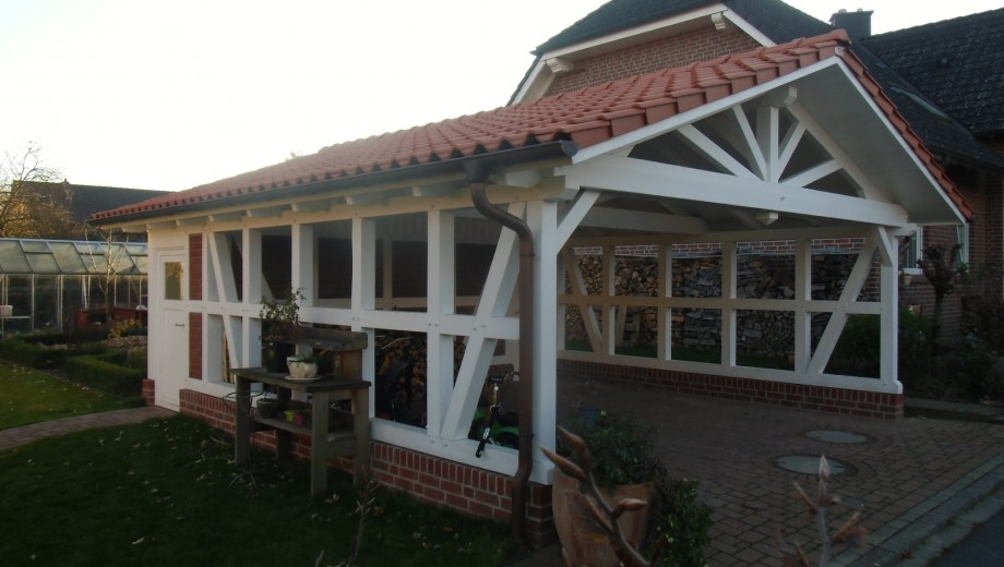 Ein Carport mit roten Ziegeln und weißen Balken neben einem Haus, umgeben von einem gepflegten Garten.