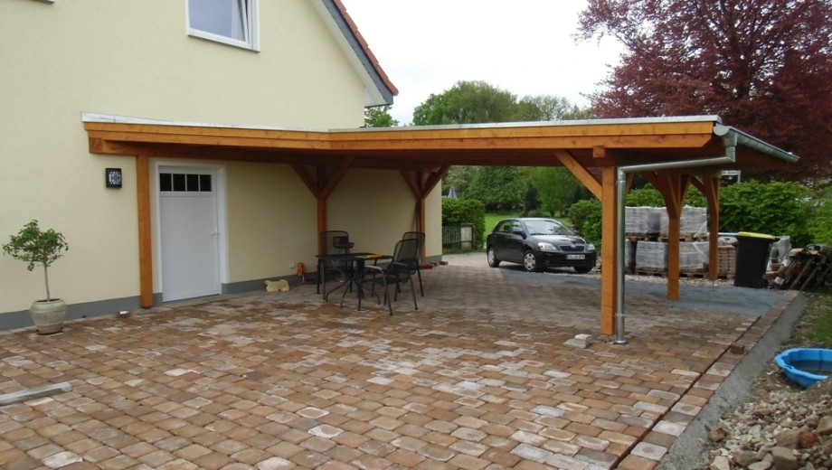 Ein Carport aus Holz an einem Haus mit geparktem Auto und Tisch auf gepflastertem Boden, umgeben von Vegetation.
