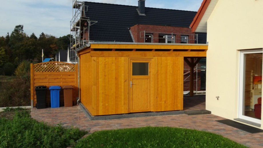 Ein Holzschuppen im Garten neben einem Wohnhaus, mit Mülleimern und einem Gebäude im Hintergrund.