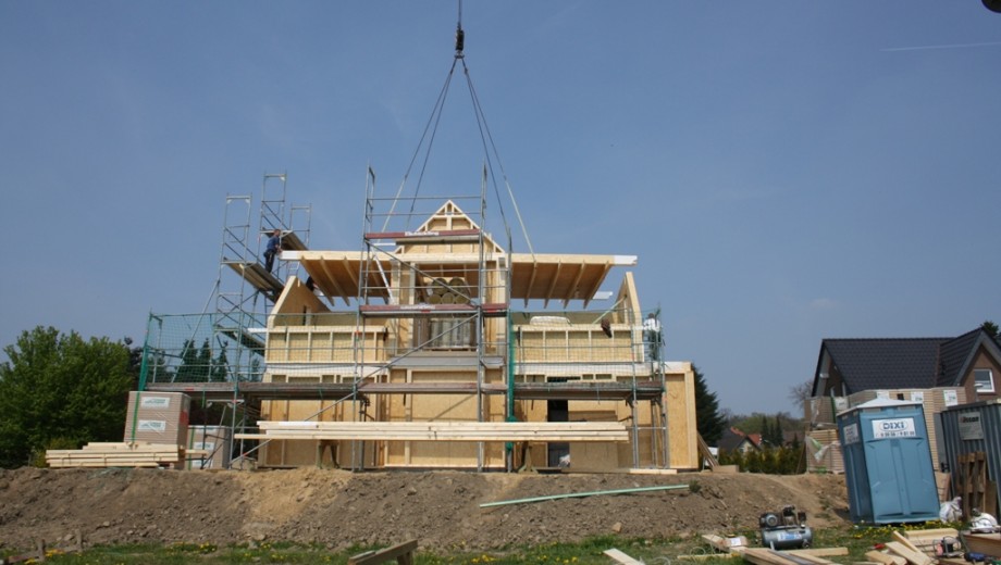 Baustelle eines Einfamilienhauses mit Kran, Baugerüst und arbeitender Person unter klarem Himmel.