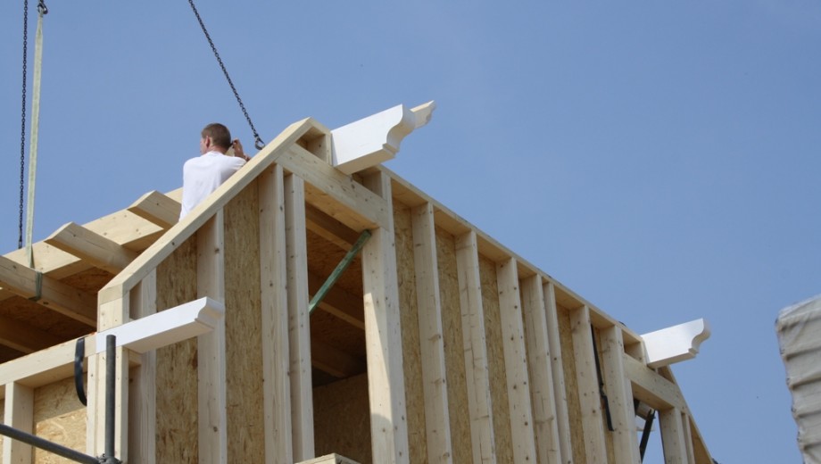 Ein Arbeiter auf einem Holzhausdach, umgeben von sichtbaren Holzbalken und einem Kran unter blauem Himmel.