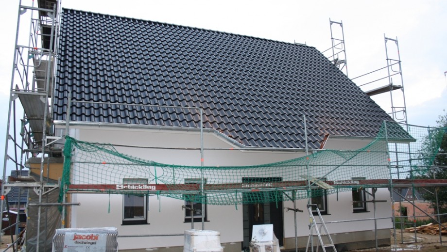 Ein im Bau befindliches Einfamilienhaus mit Gerüst und Schutznetzen, umgeben von Baumaterialien.