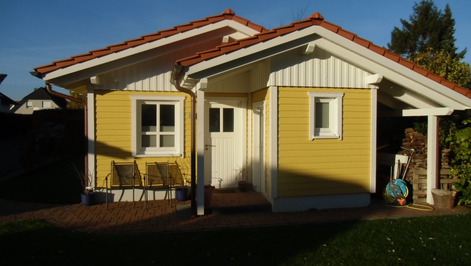 Ein gelbes Haus mit rotem Ziegeldach, umgeben von Gartenstühlen und Topfpflanzen, strahlt Gemütlichkeit aus.