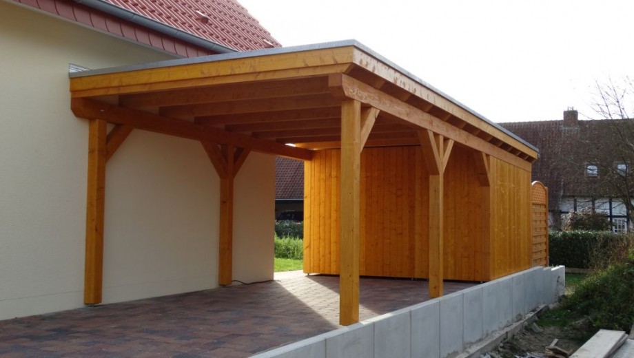 Ein hölzerner Carport mit Flachdach ist an ein helles Gebäude mit rotem Ziegeldach angebaut.