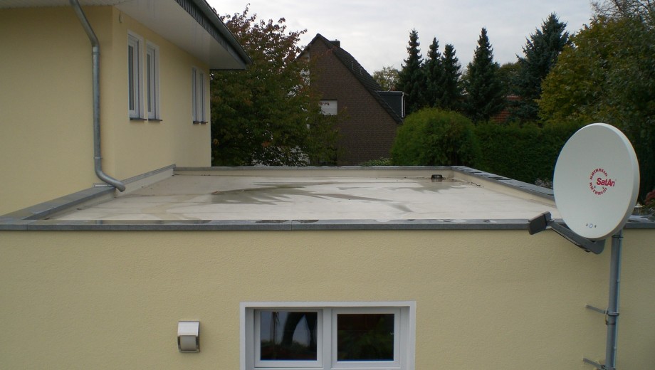 Ein Flachdachgebäude mit Satellitenschüssel, Fenster und Bäumen im Hintergrund an einem bewölkten Tag.