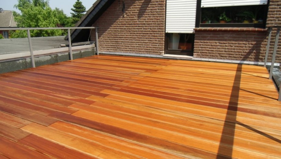 Holzterrasse auf einem Dach mit Ziegelgebäude im Hintergrund, umgeben von einem Metallgeländer.