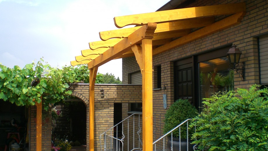 Eine gelbe Holzpergola an einem Backsteingebäude mit grüner Vegetation, die eine einladende Atmosphäre schafft.