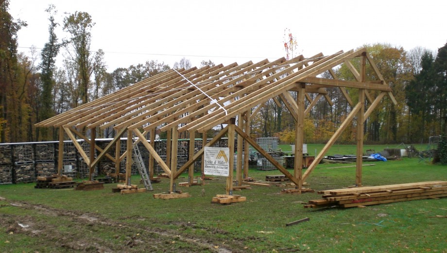 Holzgerüst auf ländlicher Baustelle, umgeben von Herbstbäumen und Baumaterialien auf feuchtem Boden.