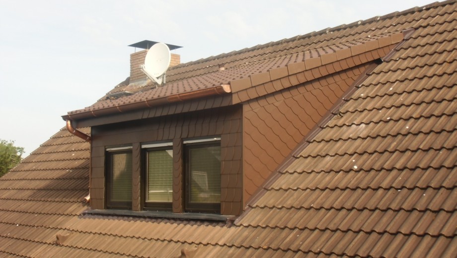 Ein braunes Dach mit Gaube und Fenstern, daneben ein Schornstein mit Satellitenschüssel unter bewölktem Himmel.