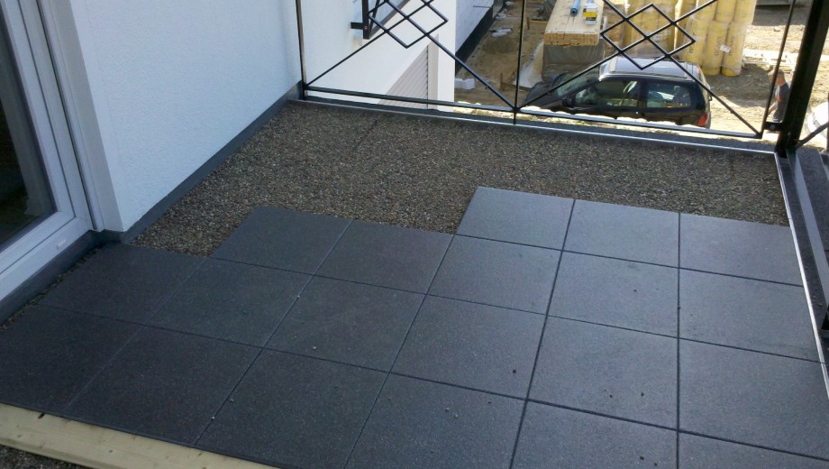 Ein Balkon mit dunklen Fliesen, Kiesfläche und schwarzem Geländer vor einem weißen Gebäude.