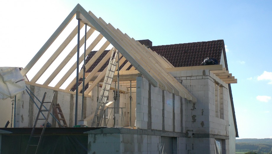 Baustelle mit Rohbau eines Hauses, sichtbare Holzträger für das Dach und mehrere Leitern. Klarer Himmel.