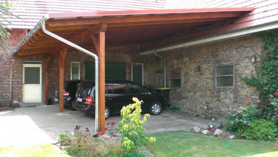 Zwei schwarze Autos parken unter einem roten Ziegel-Carport neben einer Natursteinwand, umgeben von Pflanzen.