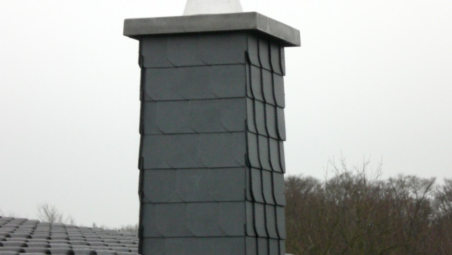 Ein rechteckiger Schornstein mit Schieferplatten auf einem dunklen Ziegeldach vor wolkigem Himmel.