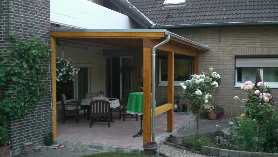 Überdachte Terrasse mit Holzkonstruktion, grüner Tischdecke, Gartenmöbeln und blühenden Pflanzen.