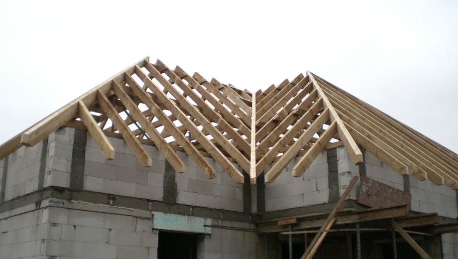 Ein Rohbau mit Dachstuhl aus Holzbalken auf Betonwänden, unterstützt von Baugerüsten unter wolkigem Himmel.