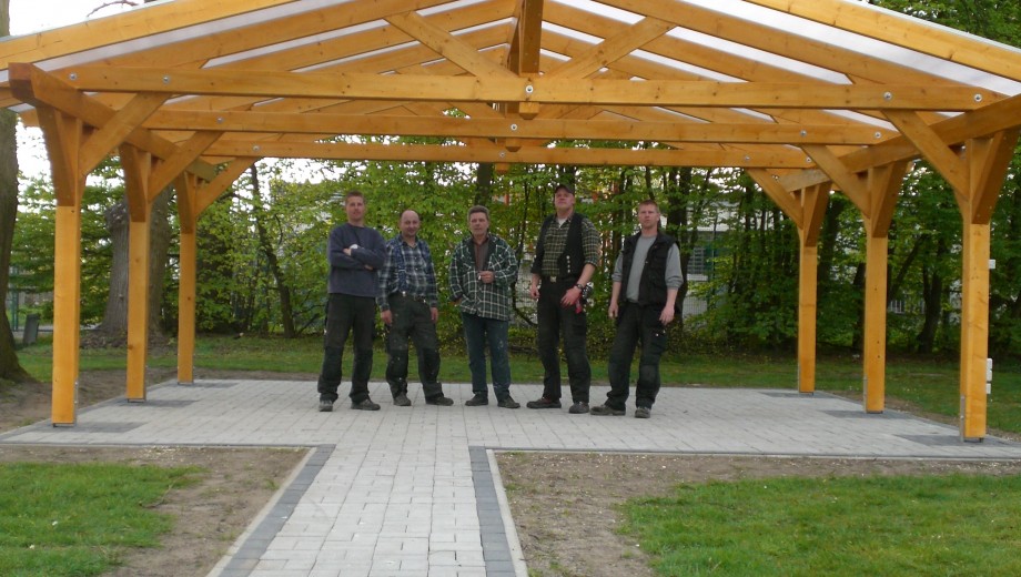 Fünf Personen posieren stolz unter einem neu errichteten Holzpavillon in einer grünen Umgebung.