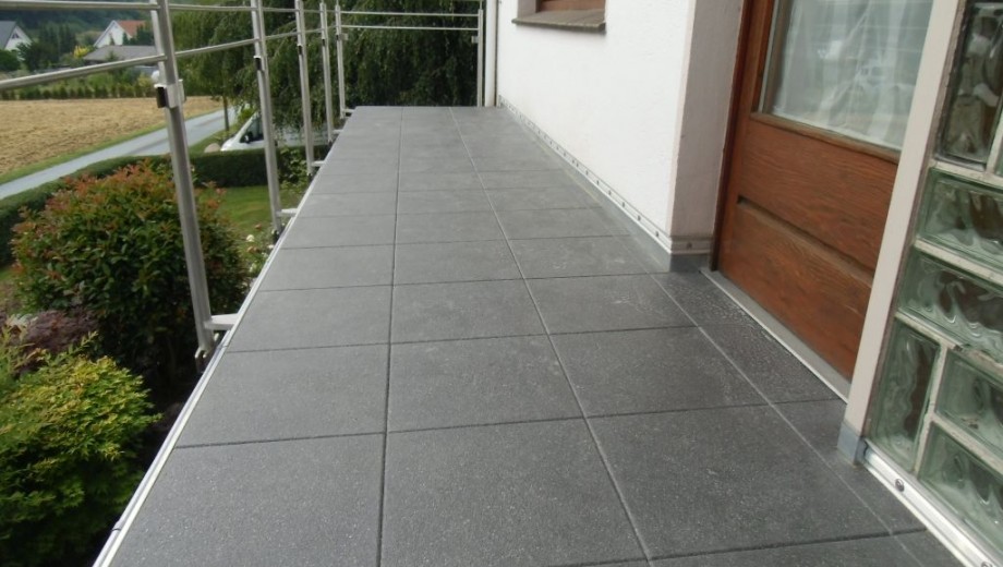 Eine gepflegte Terrasse mit dunkelgrauen Fliesen, weißer Hauswand und einer braunen Holztür.