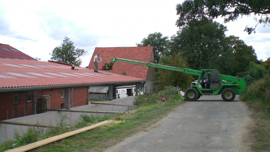 Ein Mann arbeitet auf einem Dach, unterstützt von einem grünen Teleskoplader in ländlicher Umgebung.