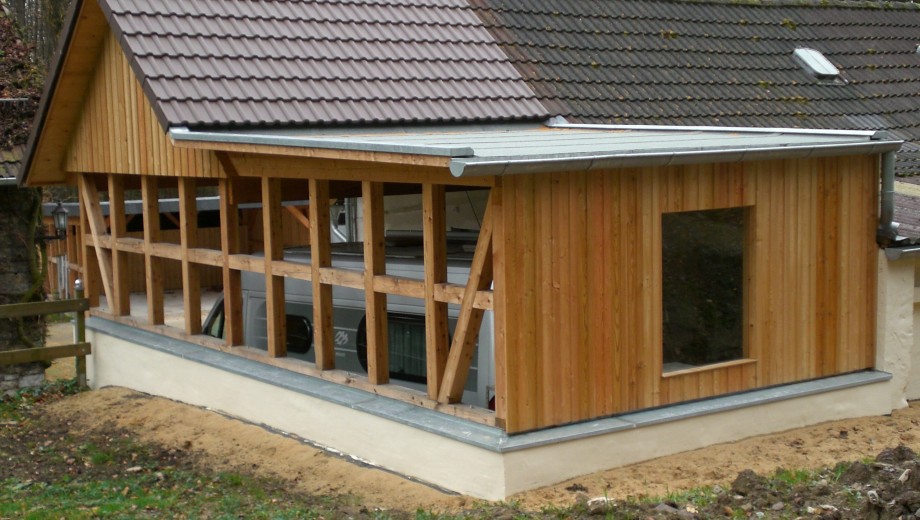 Ein Holzcarport mit geneigtem Dach, teilweise ein Auto sichtbar, umgeben von erdigem Boden.