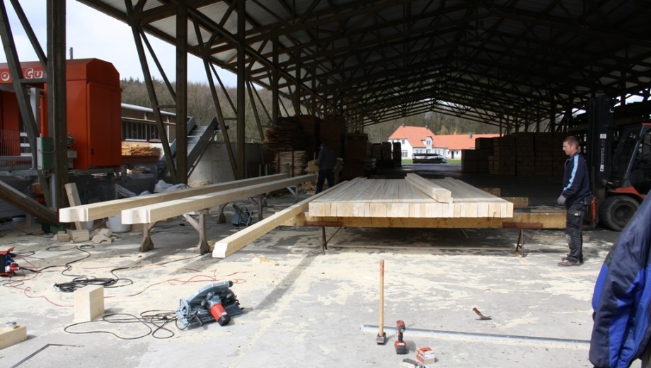 Eine geschäftige Holzverarbeitungsanlage mit Arbeitern, Gabelstapler und großen Holzbalken.