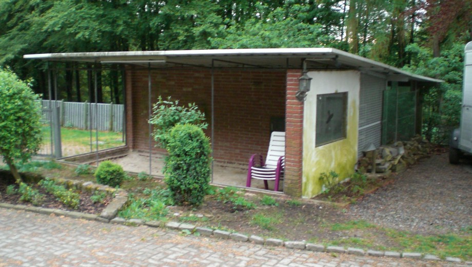 Ein einfacher Schuppen aus Ziegeln und Wellblech, umgeben von grüner Vegetation, mit gestapelten Stühlen davor.