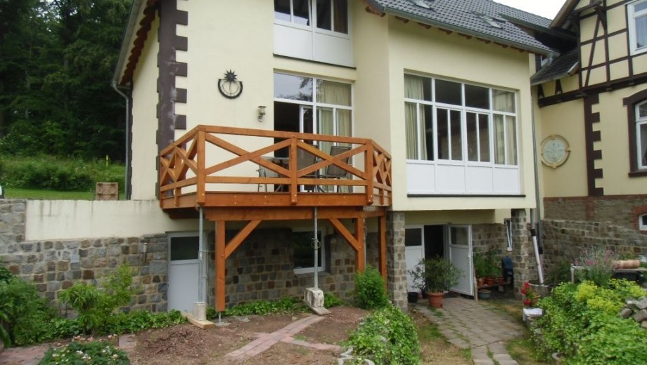 Ein zweistöckiges Haus mit hellem Holz-Balkon und großen Fenstern, umgeben von grünem Garten.