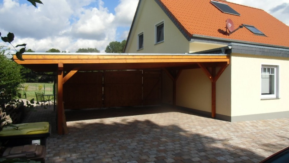 Ein Holz-Carport neben einem Einfamilienhaus mit rotem Dach und gelber Fassade, umgeben von Gartenmöbeln.