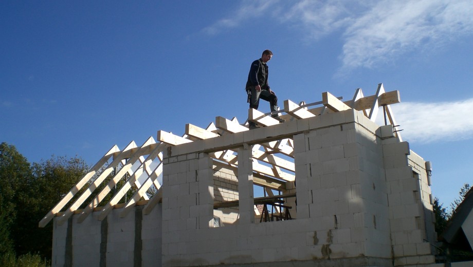 Ein Mann arbeitet auf dem Dachstuhl eines im Bau befindlichen Hauses unter einem klaren, blauen Himmel.