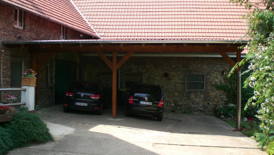 Ein hölzerner Carport mit zwei schwarzen Autos steht neben einem Gebäude mit Ziegel- und Natursteinwänden.