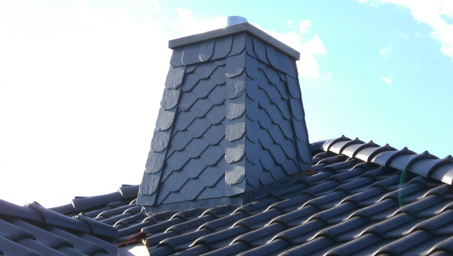 Ein Schornstein mit dunkelgrauen Schieferplatten auf einem Dach mit gleichfarbigen Ziegeln unter blauem Himmel.
