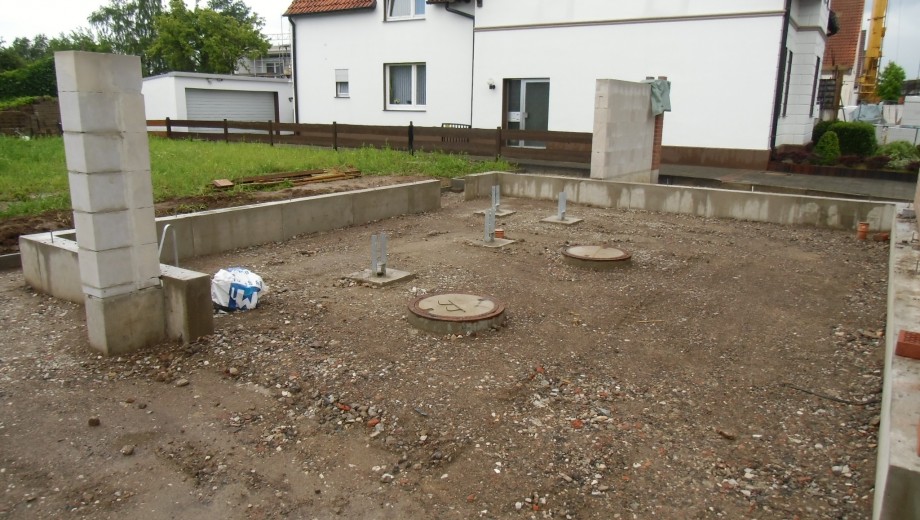 Eine Baustelle mit Betonpfeilern und Abdeckungen, im Hintergrund ein weißes Haus mit rotem Dach.