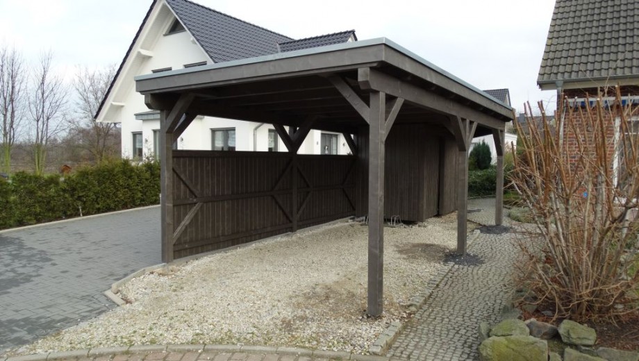 Ein dunkler Holz-Carport steht auf Kies, neben einer gepflasterten Einfahrt, vor zwei Häusern.
