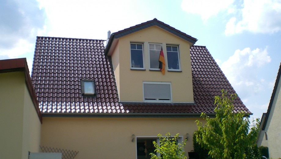 Ein zweistöckiges Haus mit rotem Dach und deutscher Flagge, umgeben von blauem Himmel und grünem Baum.
