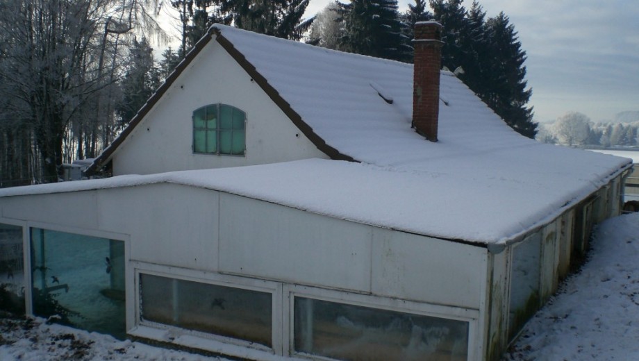 Winterliches Haus mit schneebedecktem Dach, großen Fenstern und verschneiten Bäumen im Hintergrund.