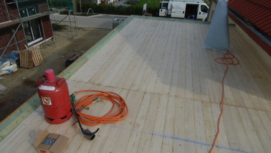 Baustelle auf Flachdach mit Gasflasche, Schläuchen, Holzplanken und Transporter im Hintergrund.
