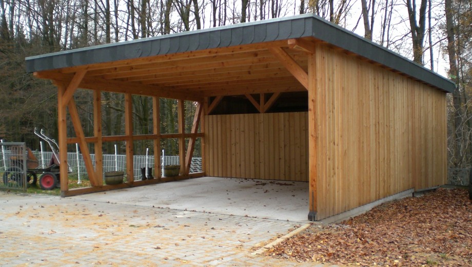 Ein hölzerner Carport in herbstlicher Umgebung mit kahlen Bäumen und Gartenwerkzeug am Rand.