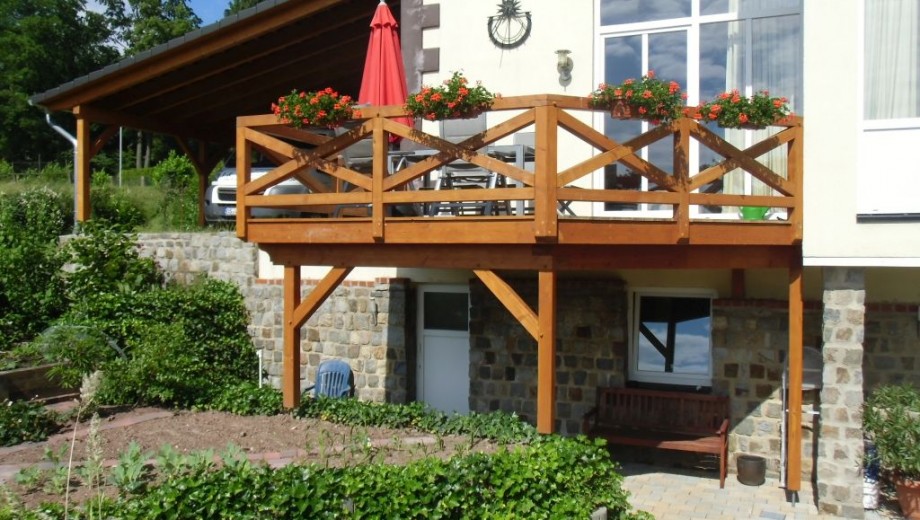 Terrasse mit Holzbalkon, Blumentöpfen, rotem Sonnenschirm; darunter Tür, Fenster, blauer Stuhl, Holzbank.