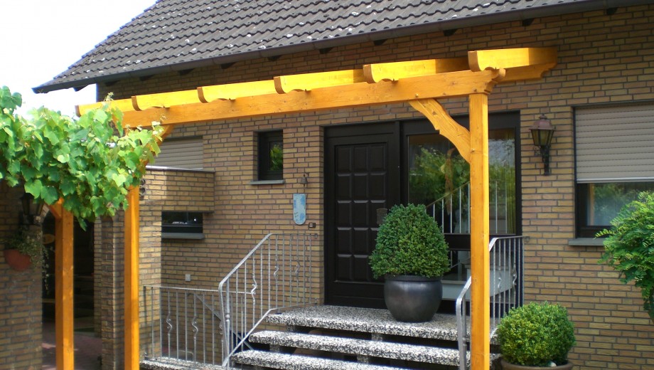 Einladender Hauseingang mit Holzpergola, Treppe, massiver Tür und flankierenden grünen Pflanzen.