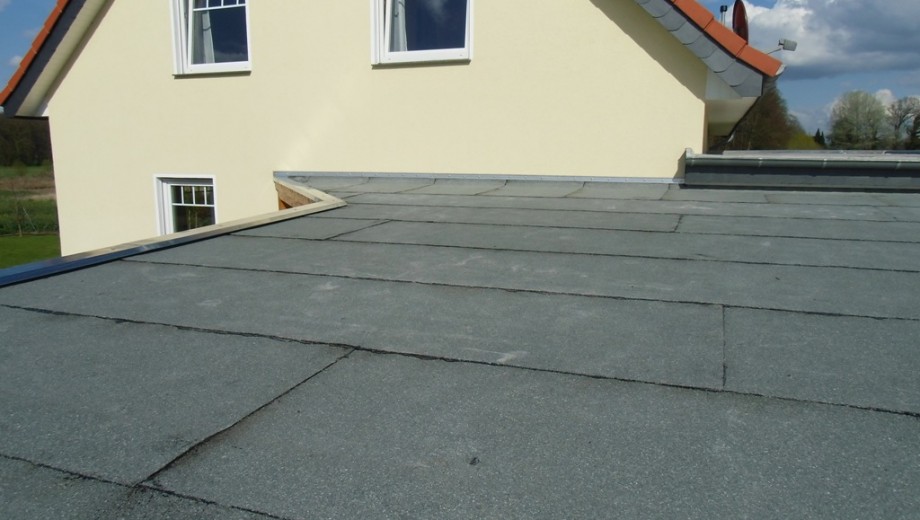 Ein Flachdach mit grauen Platten, ein Haus mit hellgelber Fassade und ein Satteldach mit orangefarbenen Ziegeln unter klarem Himmel.