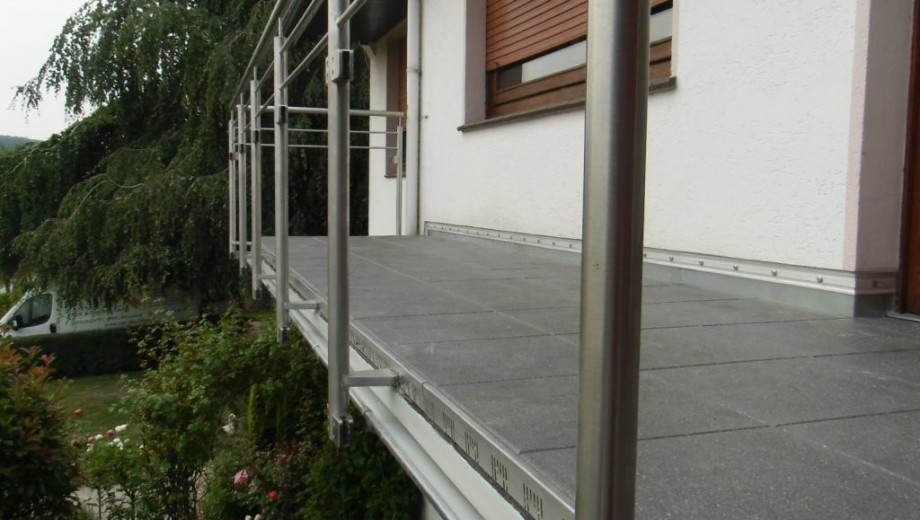 Eine ruhige Terrasse mit grauen Fliesen, silbernem Geländer, weißer Fassade und grünem Baum im Hintergrund.
