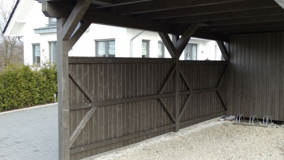 Dunkler Holz-Carport mit hellem Kiesboden, neben modernem weißem Haus in ruhiger Umgebung.