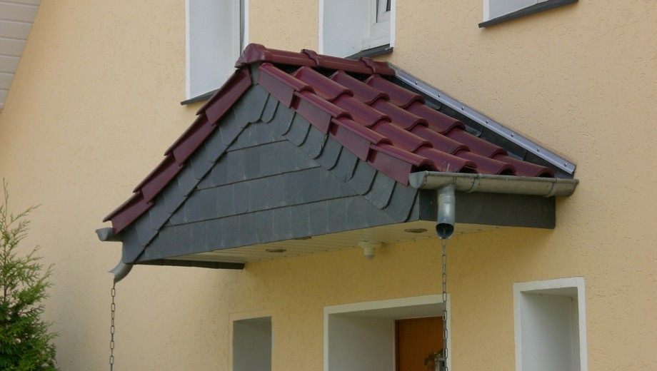Eingangsbereich mit rotem Ziegeldachvordach, gelber Fassade und metallischer Regenrinne.