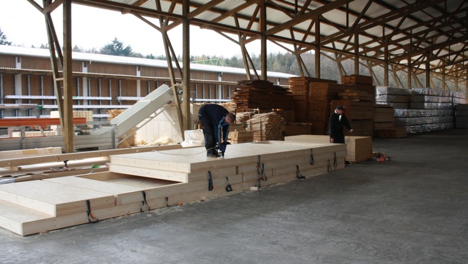 Zwei Personen arbeiten an großen Holzplatten in einer industriellen Lagerhalle.